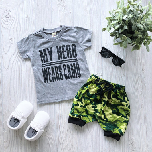 A flat lay of a childs outfit: the My Hero Wears Camo • Gray Tee, green camo shorts, white slip-on shoes, black sunglasses, and a potted plant on a light wooden surface.