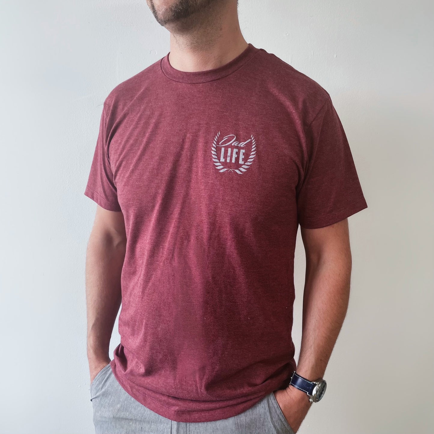 A man wears the Dad Life Crest • Maroon Tee, a unisex maroon Tultex shirt featuring Dad Life inside a laurel wreath on the chest. He stands against a plain white wall with his hands in his pockets.