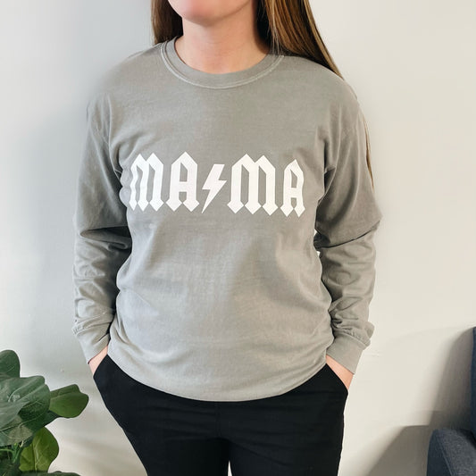 A person stands indoors with hands in pockets, wearing the MAMA Rocker • Gray Long Sleeve, a garment-dyed light gray tee featuring bold white rock-style MAMA lettering. Part of a green plant is visible on the left.