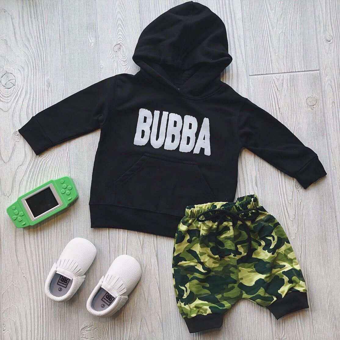 A flat lay from mini’s wardrobe: the Bubba Hoodie • Black, green camo shorts, white slip-on shoes, and a green handheld game console, all arranged on light wood flooring.