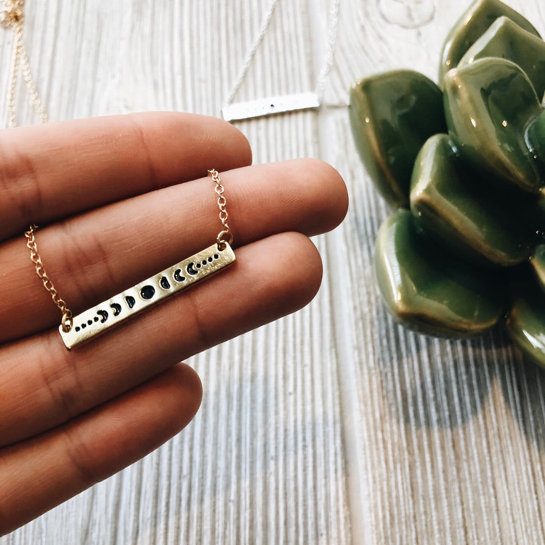 A hand displays the Moon Phase • Bar Necklace with a gold bar pendant and adjustable clasp, shown next to a green ceramic succulent and another silver or gold necklace on a light wooden surface.