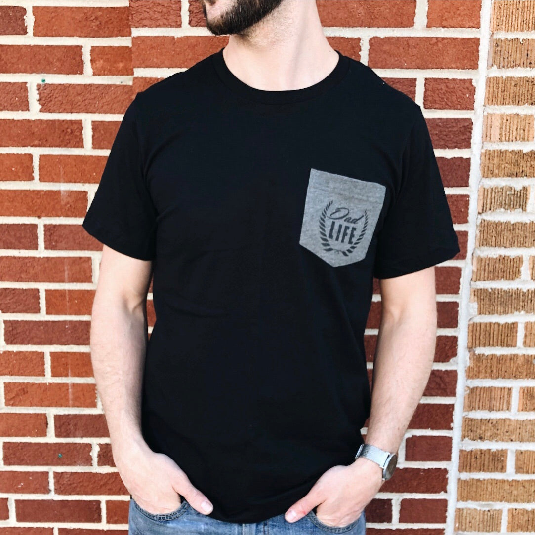 A man stands against a brick wall wearing the Dad Life • Pocket Tee, a black unisex Bella Canvas shirt with a gray pocket featuring Dad Life and a laurel graphic. Made from combed and ring-spun cotton, it pairs perfectly with jeans.