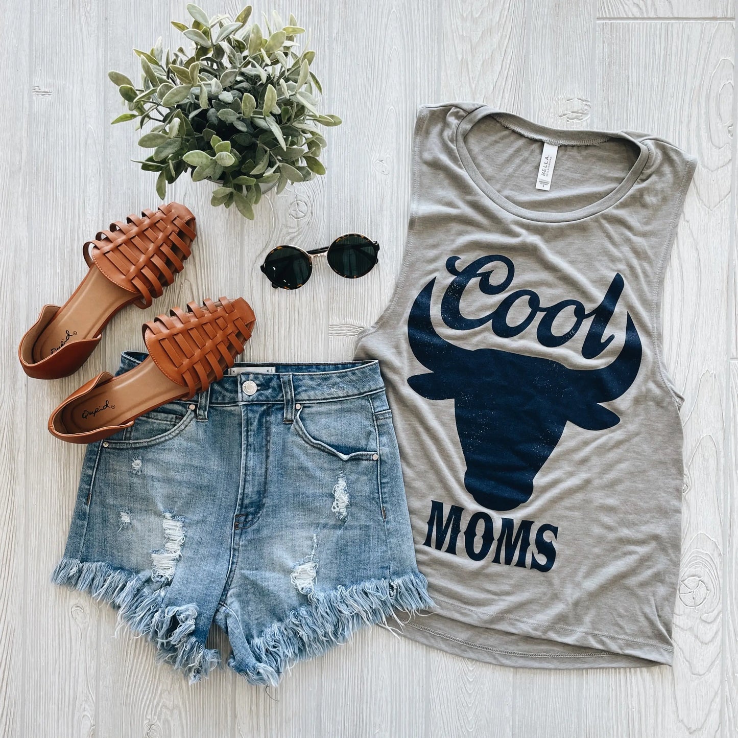 Flat lay of a casual summer outfit featuring the Cool Moms • Stone Scoop Tank with a bull graphic, distressed denim shorts, brown sandals, black round sunglasses, and a small potted plant.