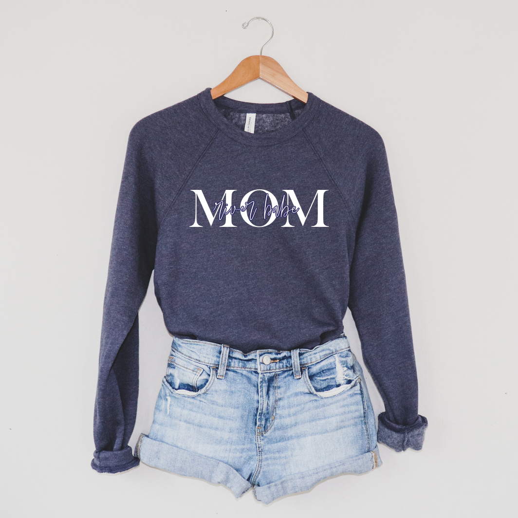 The MOM River Babe • Navy Pullover, featuring “MOM” in bold white letters and “mama bear” script across the chest, hangs on a wooden hanger above light denim shorts, showcasing its cozy unisex fit against a plain wall.