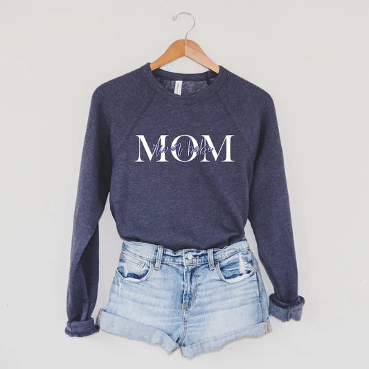 The MOM River Babe • Navy Pullover, featuring “MOM” in bold white letters and “mama bear” script across the chest, hangs on a wooden hanger above light denim shorts, showcasing its cozy unisex fit against a plain wall.