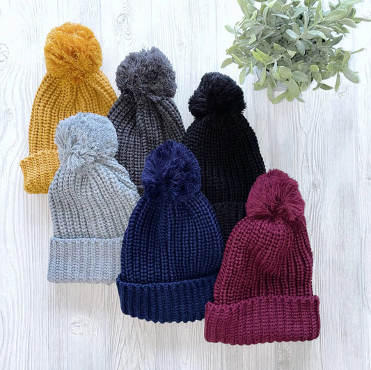 Six Chunky Ribbed Pom Beanies in yellow, grey, black, light blue, navy, and burgundy are displayed on a white wooden surface beside a small green plant.