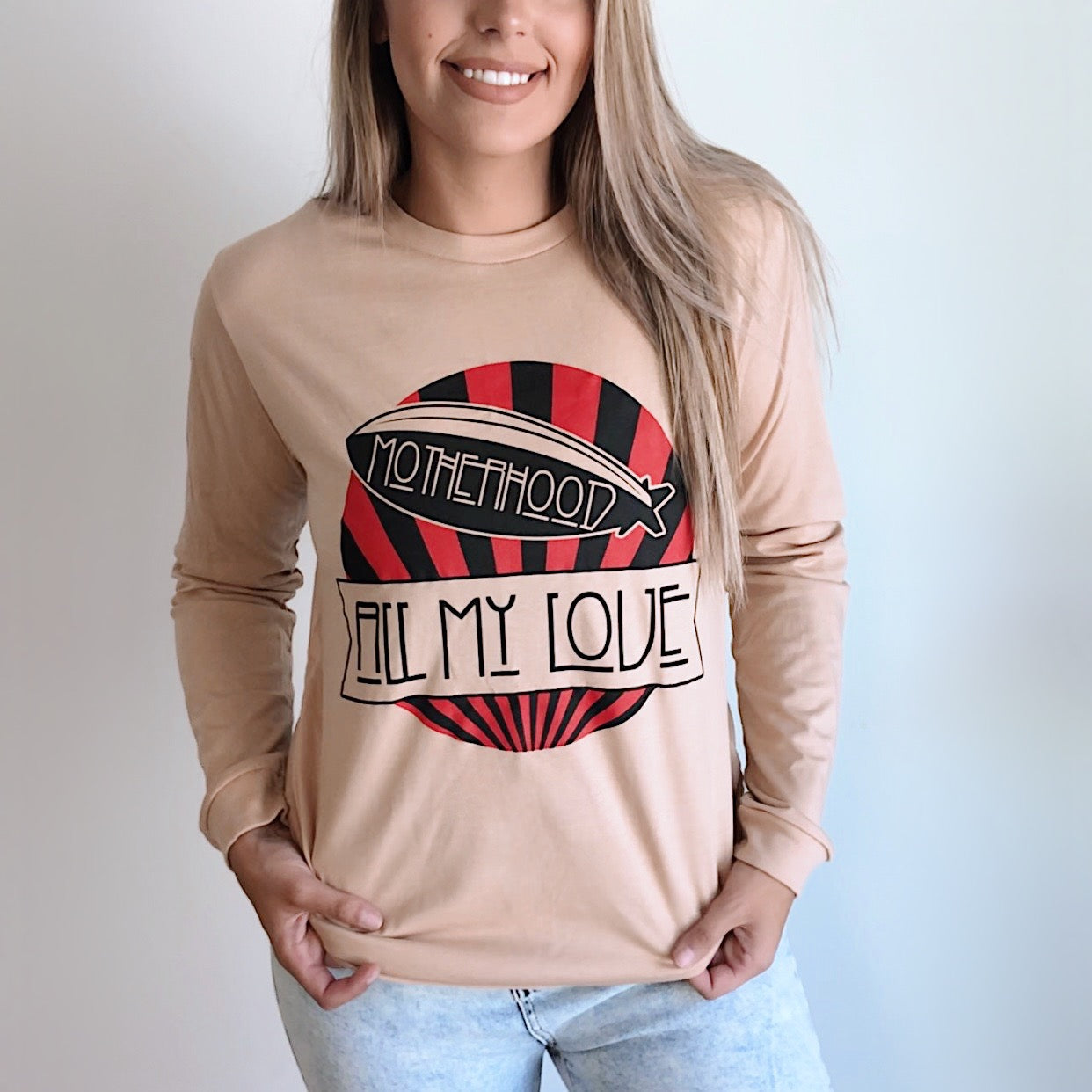 A person wears the All My Love • Long Sleeved tee, a beige unisex shirt with a blimp graphic and MOTHERHOOD ALL MY LOVE in red, black, and white. Smiling, they tuck one hand into their jeans pocket.