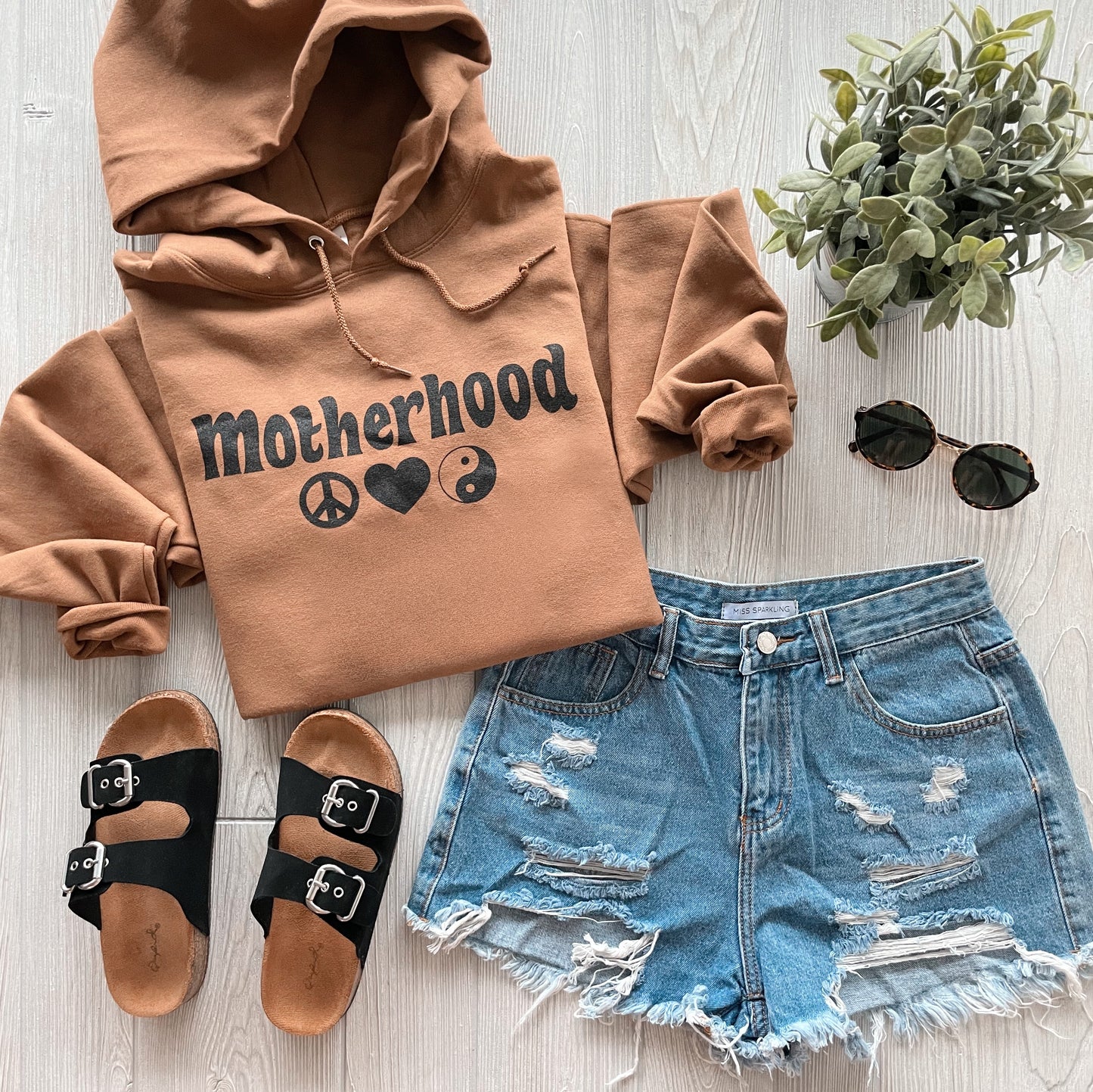 Flat lay of the Peace & Love • Caramel Hoodie featuring peace, heart, and yin-yang symbols, styled with distressed denim shorts, black sandals, round sunglasses, and a potted plant—crafted from pill-resistant fleece for lasting comfort.