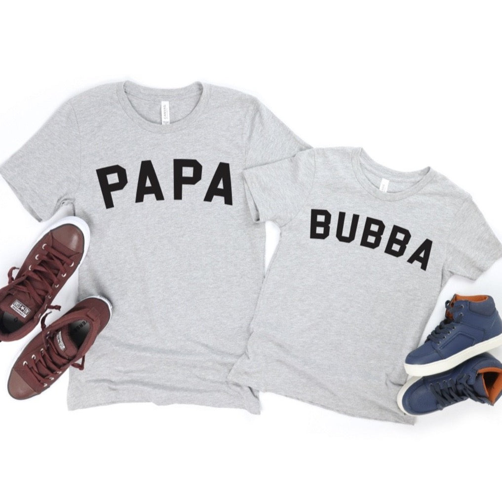 Two gray unisex New PAPA • Tees with bold black PAPA and BUBBA text are displayed beside red and blue sneakers on a white background, crafted from soft combed and ring-spun cotton for all-day comfort.