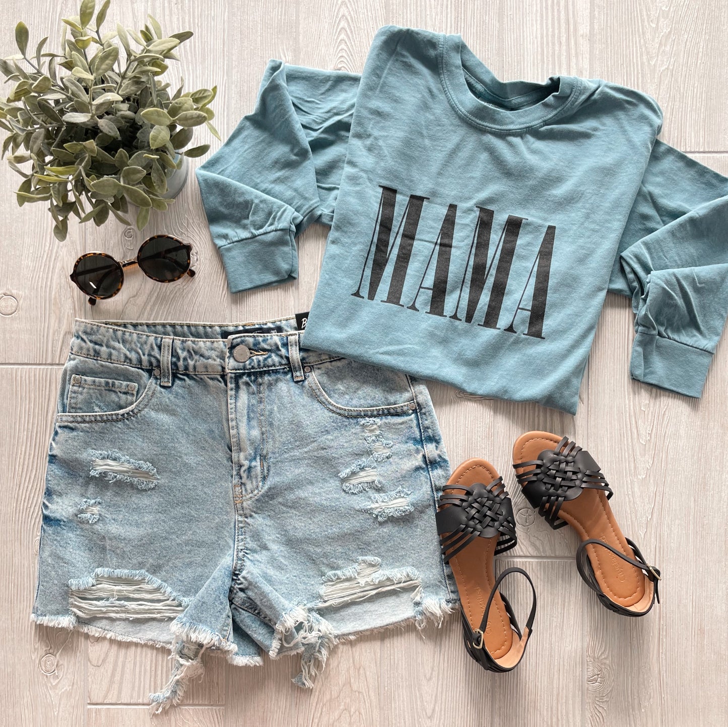 A flat lay featuring the MAMA • Ice Blue Long Sleeve made from soft ring-spun cotton, paired with distressed denim shorts, black sandals, round sunglasses, and a small potted plant on a light wooden surface.