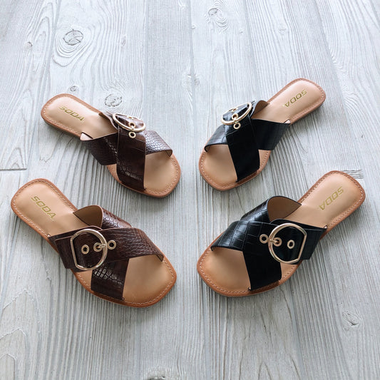 Two pairs of Buckle Sandal • Cognac or Black! are shown on a light wood floor. Both have crisscross straps with gold buckles and SODA on the insole—one pair in cognac faux crocodile print, the other in black.