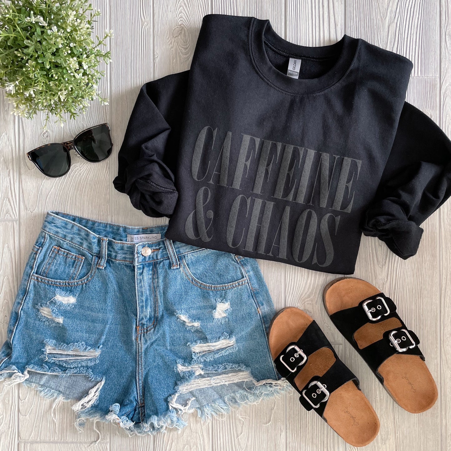 The Caffeine and Chaos • Tonal Pullover in black is styled with distressed denim shorts, black buckle sandals, black sunglasses, and a small potted plant, all displayed on a light wood surface.