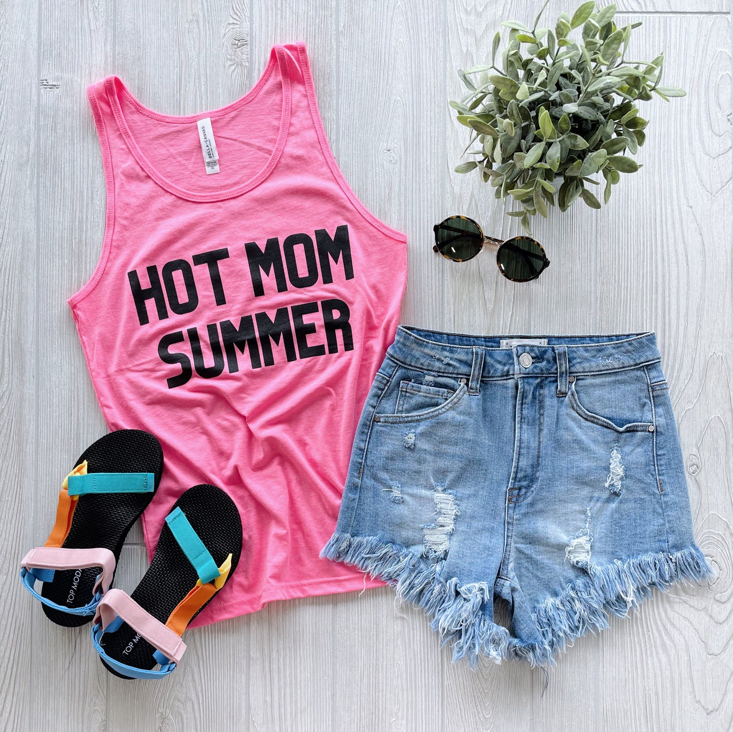 A Hot Mom Summer • Unisex Tank Top in hot pink is styled with distressed denim shorts, black sunglasses, colorful sandals, and a potted plant on a light wood surface.