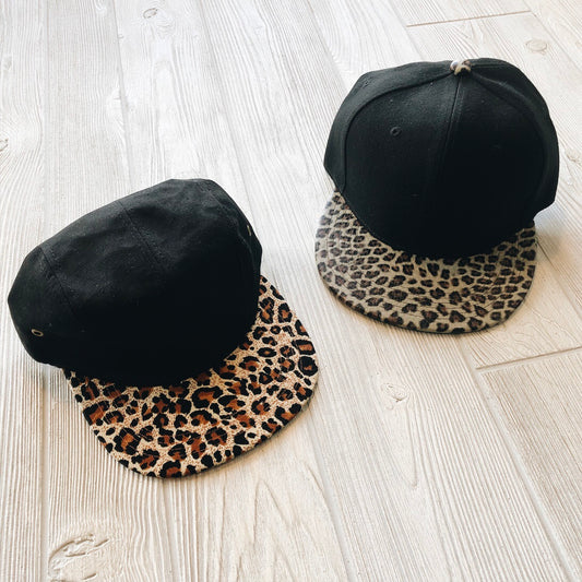 Two black Mommy & Me • Cheetah Hat caps sit on a light wooden floor. The adult version has a brown and black leopard brim; the child’s has gray and black, both featuring adjustable straps for a perfect fit.