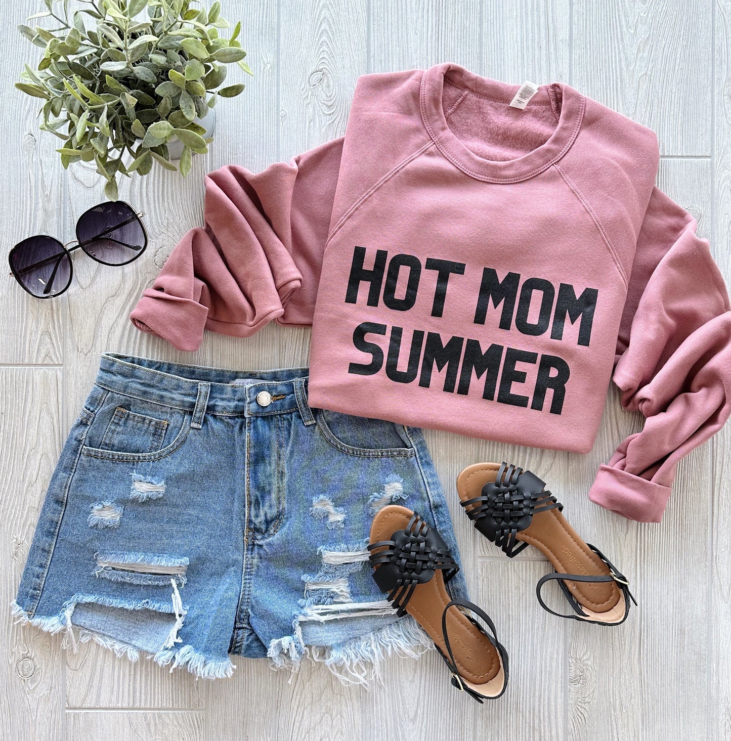 Flat lay of the Hot Mom Summer • Mauve Pullover with a unisex fit, styled with distressed denim shorts, black fringe sandals, black sunglasses, and a small green plant on a light wooden background.