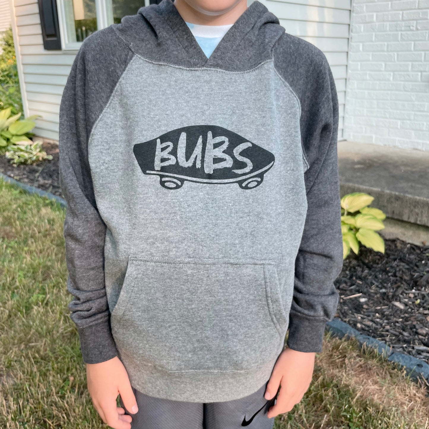 A child’s torso is shown outdoors in the Bubs • Block Sleeve Hoodie, featuring a skater graphic and “BUBS” on the front, with shrubs and a house in the background.