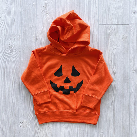 The Jack O Lantern • Orange Hoodie for kids features a black jack-o-lantern face on the front and is shown laid flat on a light wood surface. Perfect for Halloween—size up for a comfy fit!.
