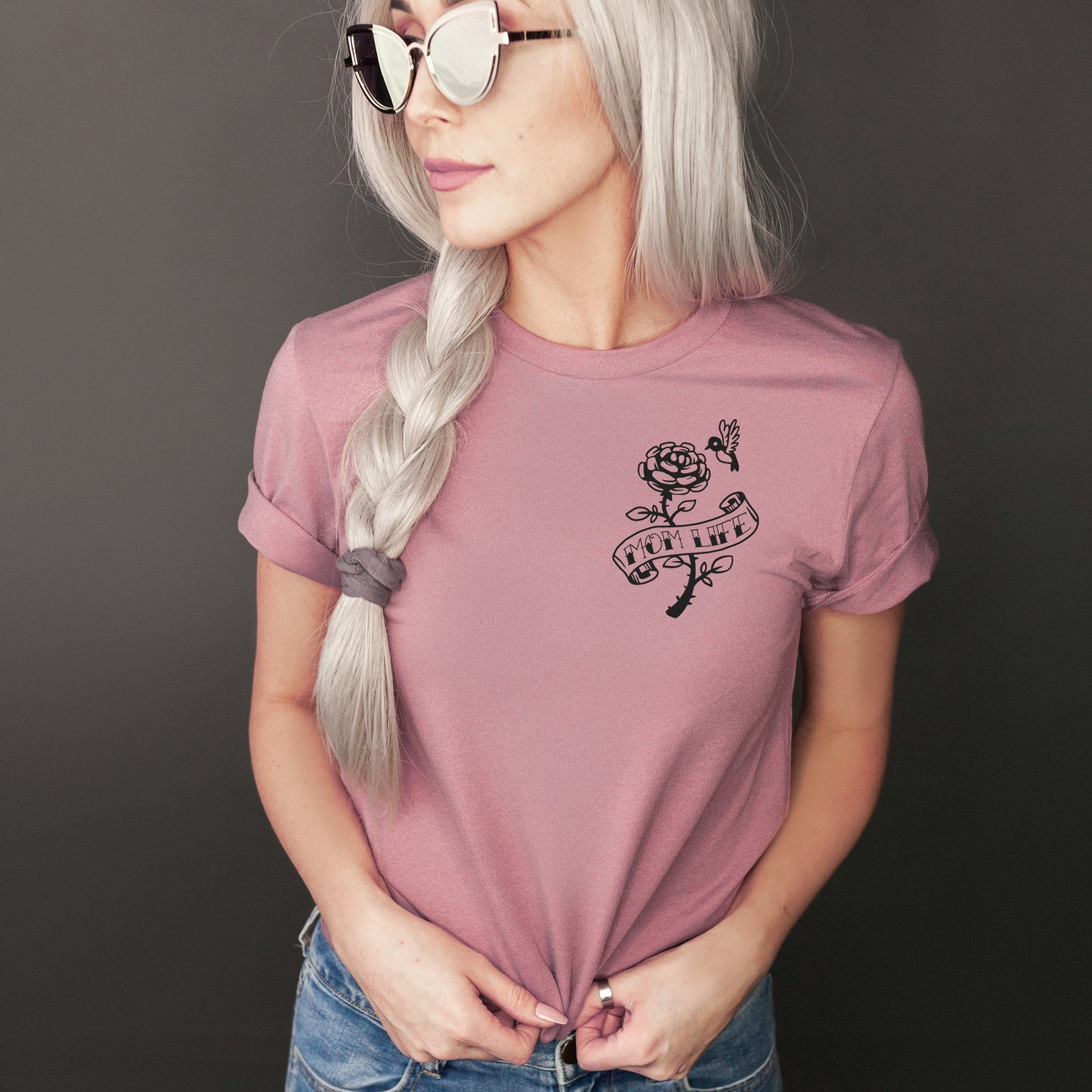 A woman with long, braided silver hair wears sunglasses and a mauve Mom Life Hummingbird • Tee with a floral banner design reading GROWTH. She looks away, holding her shirt against a dark background.