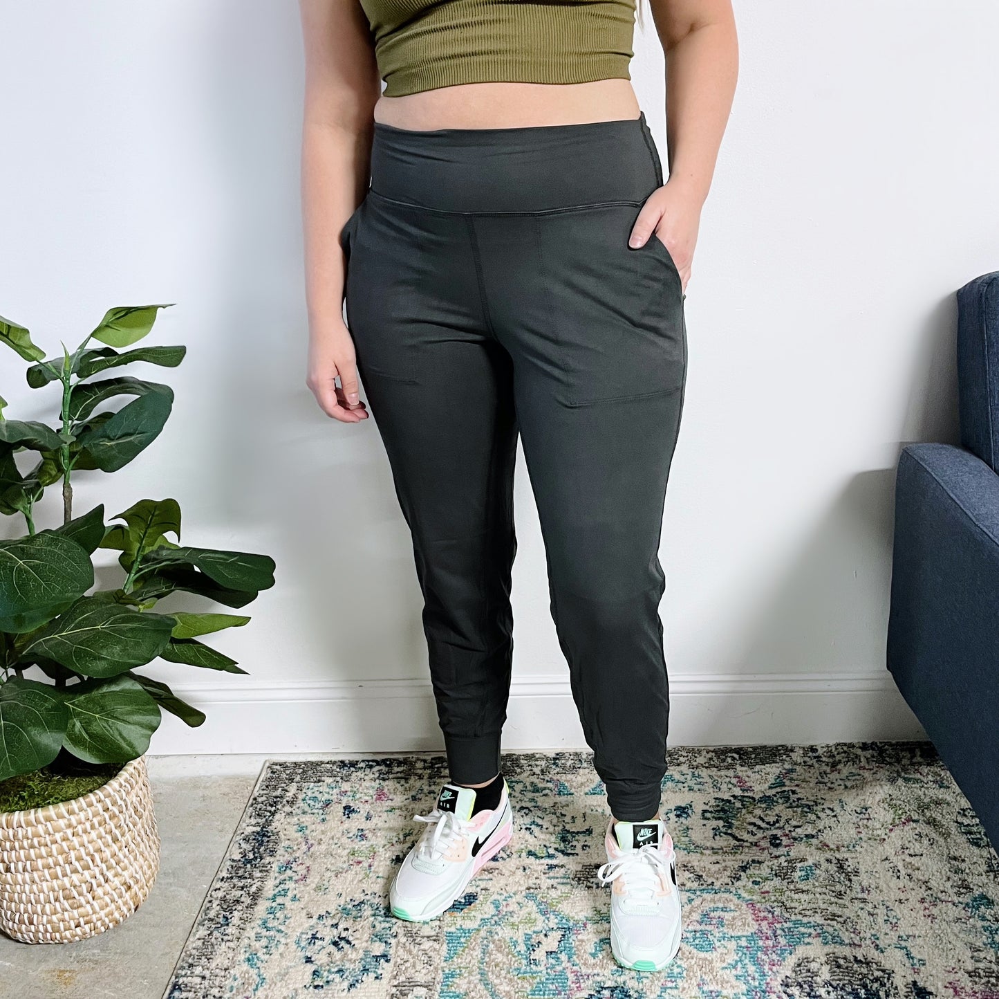 Wearing the Lexi Yoga Pant in Charcoal, a person stands indoors on a patterned rug near a potted plant and blue sofa. Theyre also dressed in a green crop top and white sneakers, with only the lower half of their face visible.