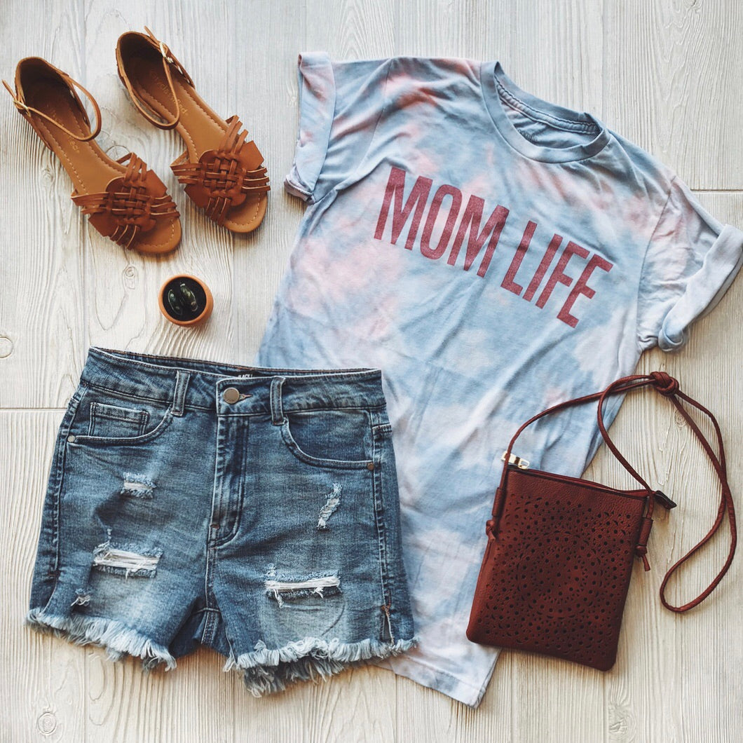 Flat lay of a casual outfit: the soft and comfy MOM LIFE • Coral Tie-Dye T-shirt, distressed denim shorts, brown sandals, a small brown crossbody bag, and a round black accessory arranged on a light wooden surface.