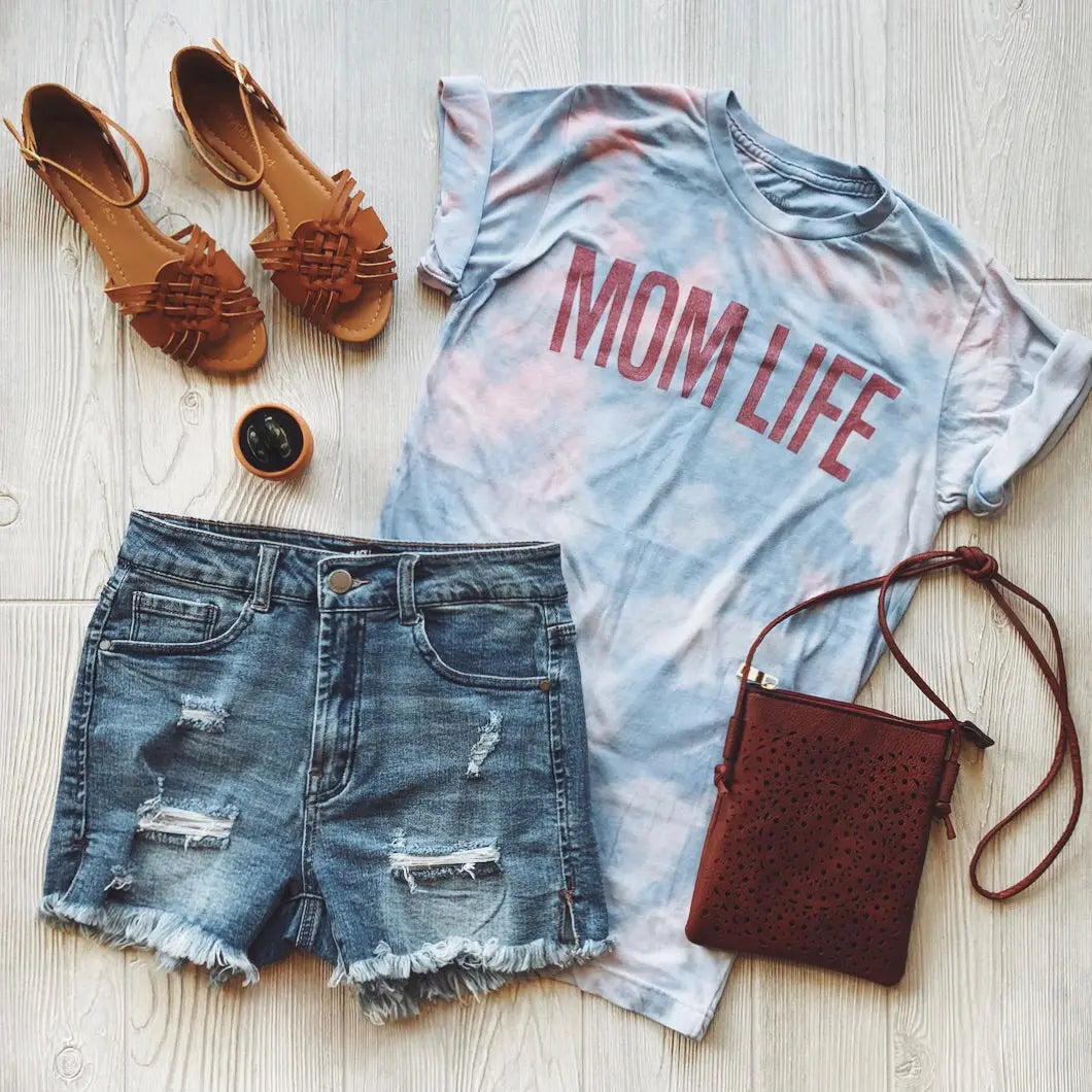 Flat lay of a casual outfit: the soft and comfy MOM LIFE • Coral Tie-Dye T-shirt, distressed denim shorts, brown sandals, a small brown crossbody bag, and a round black accessory arranged on a light wooden surface.