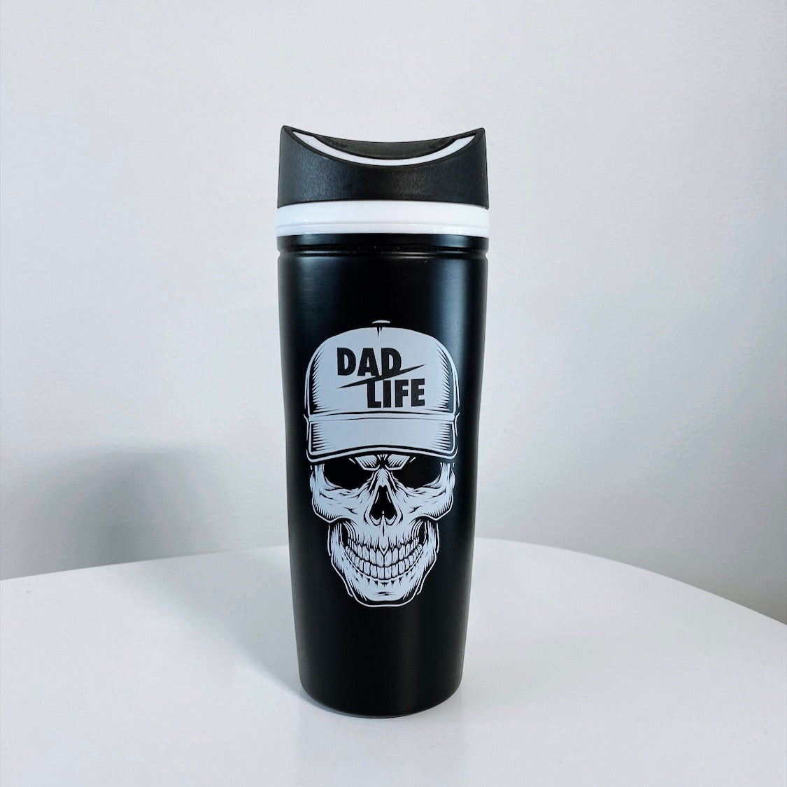 A black stainless steel Dad Skull Tumbler • Slightly Flawed, featuring a skull print with a cap that reads "DAD LIFE" in bold white letters, sits on a white surface against a plain light background.