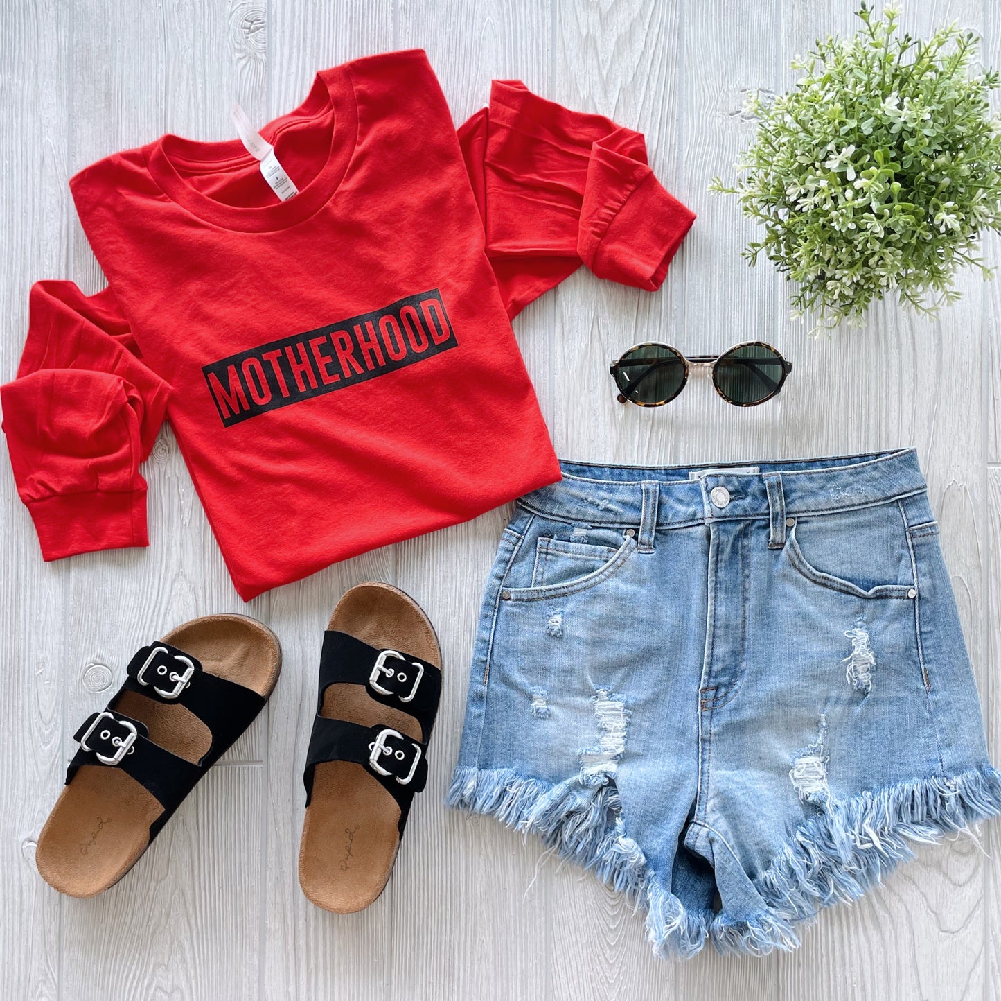 A flat lay photo of the MOTHERHOOD • Poppy Long Sleeve, paired with distressed denim shorts, black sandals, round sunglasses, and a small potted plant on light wood—perfect for highlighting cozy motherhood style.