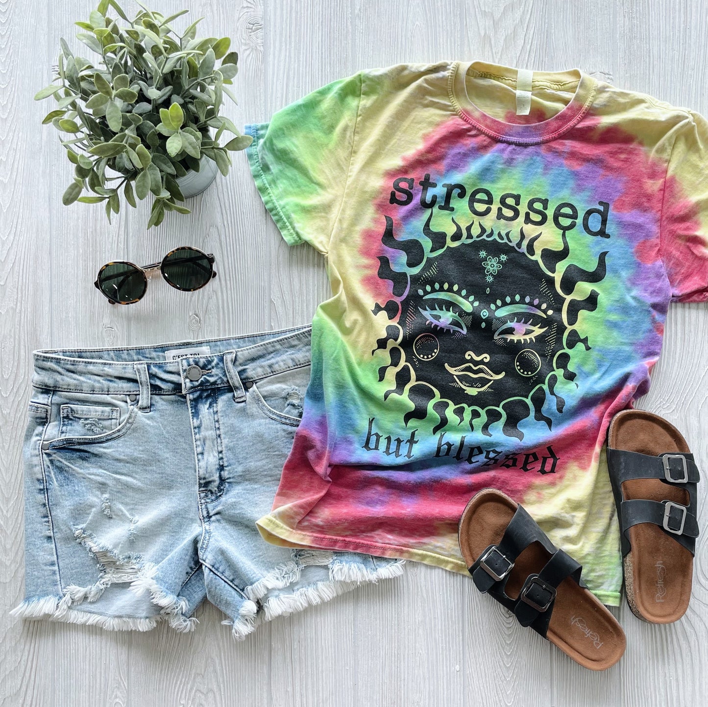 Flat lay of a casual summer outfit: Stressed • Tie Dye Tee with a sun face, denim shorts, black sandals, round sunglasses, and a small potted plant on light wood—a comfy mom tee ideal for relaxed days.