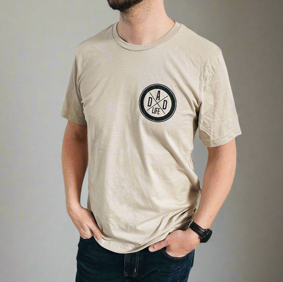 A man wears the Dad Life Left Chest • Tan Tee, featuring a black DAD LIFE print with two crossed arrows on the left chest. The beige Bella Canvas shirt offers unisex sizing for a comfortable fit, perfect for everyday style.