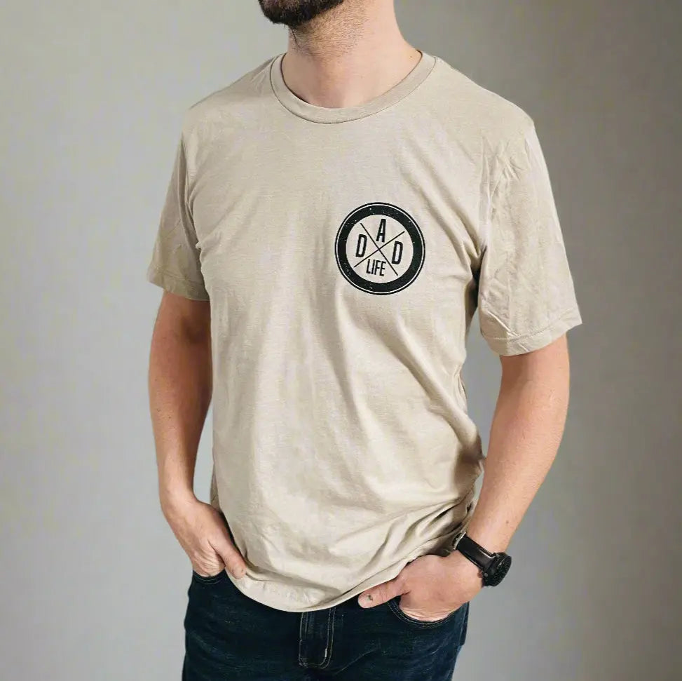 A man wears the Dad Life Left Chest • Tan Tee, featuring a black DAD LIFE print with two crossed arrows on the left chest. The beige Bella Canvas shirt offers unisex sizing for a comfortable fit, perfect for everyday style.