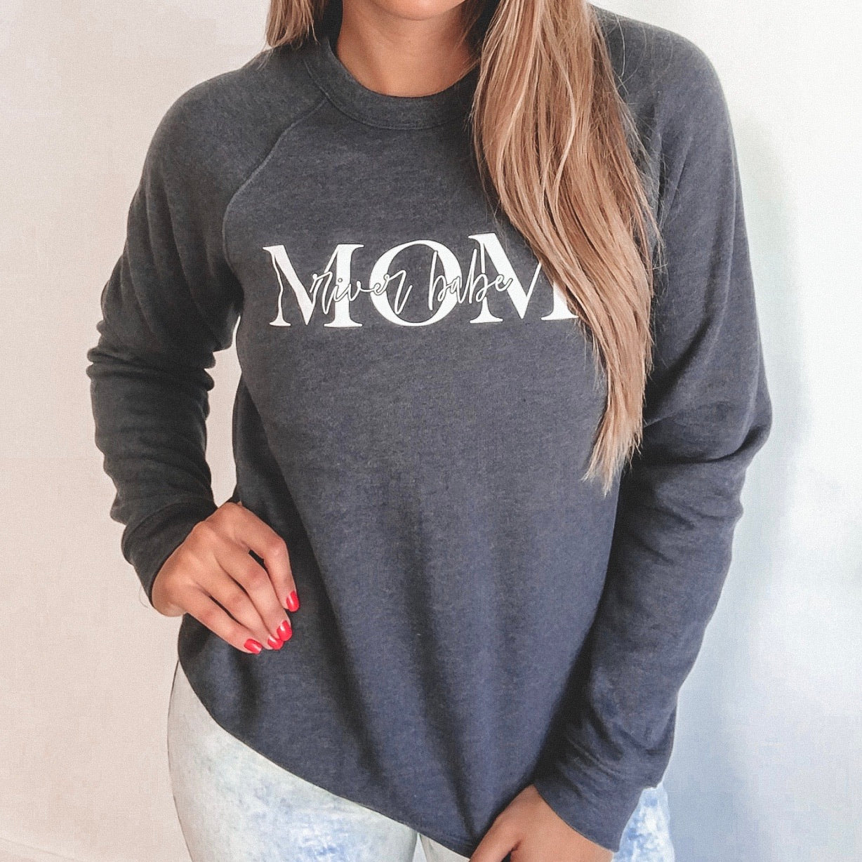 A woman with long blonde hair wears the MOM River Babe Navy Pullover, a unisex fit sweatshirt featuring MOM in large white letters with mini babe in script above, paired with light-colored pants and one hand on her hip.