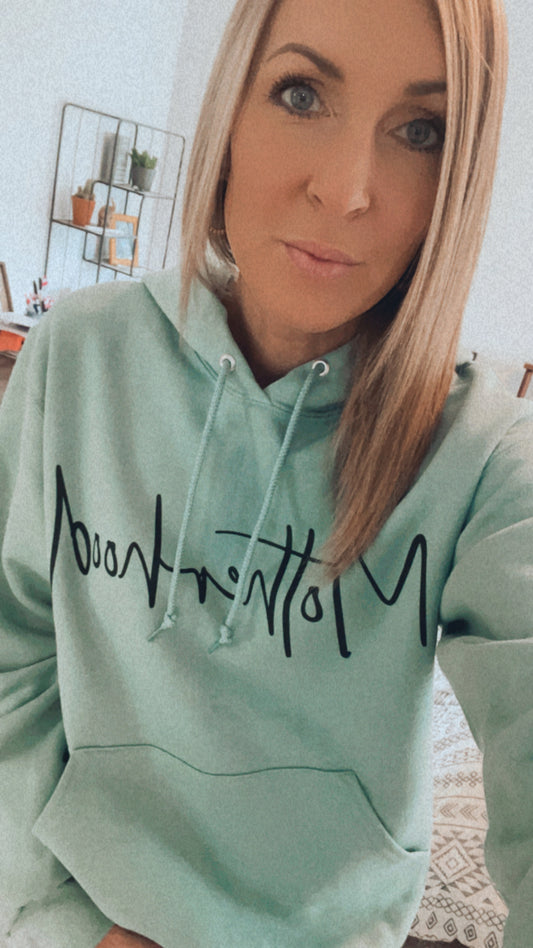A woman with straight, blonde hair takes an indoor selfie in a Motherhood • Sage Hoodie featuring black text. The unisex fit stands out as she poses, with shelves filled with plants and decor visible in the background.