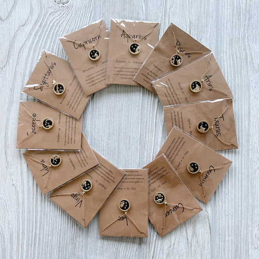 A flat lay of twelve brown paper envelopes arranged in a circle, each labeled with a zodiac sign and containing a small moon-and-stars pin—an ideal pairing for your Zodiac Sign • Necklace—displayed on a light wooden surface.