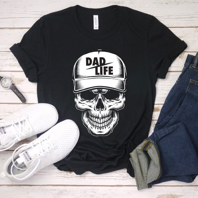 The Skeleton Dad Life Tee is a black Bella Canvas unisex essential t-shirt featuring a skull in a baseball cap with DAD LIFE text. Made from soft combed and ring-spun cotton, its styled with white sneakers, a watch, and blue jeans on wood.