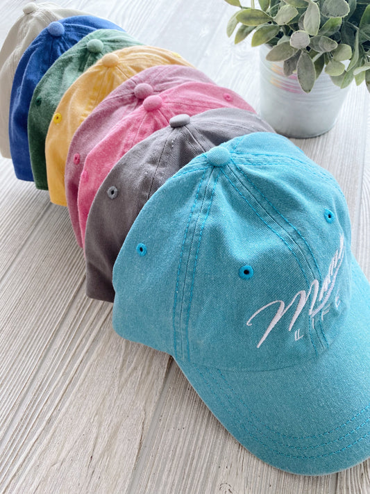Six Mama Life Hat • NEW Colors! caps in blue, green, yellow, red, gray, and turquoise are arranged in a row on a light wooden surface next to a potted plant.