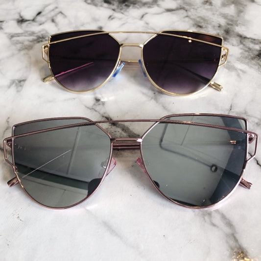 Two pairs of Claire UV Protected sunglasses with metal frames rest on marble—one gold with dark lenses, the other rose gold with grayish lenses. Both are ready to ship and available in multiple colorways for adults!.