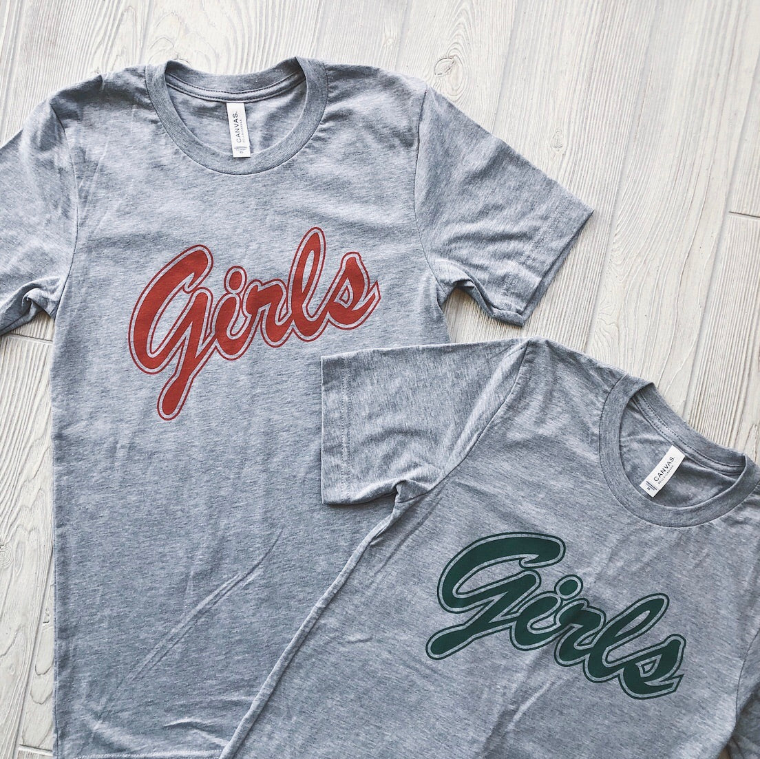 Two gray Girls • Tees are displayed on a light wood floor. Each shirt says Girls in large script—one in red, one in green—creating a playful, Friends-inspired look.