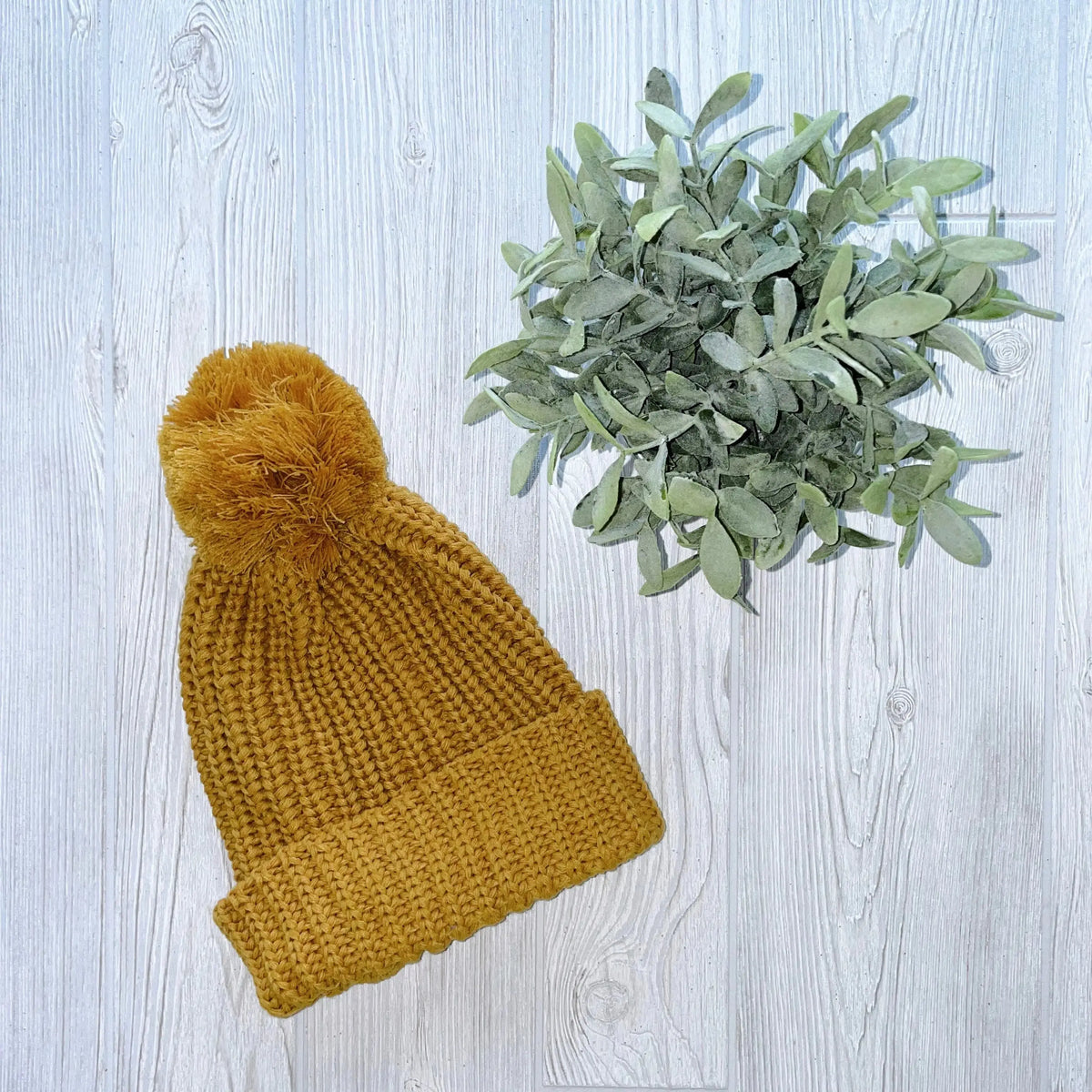 A Chunky Ribbed Pom Beanie in mustard yellow sits beside a small potted green plant on a light wooden surface.