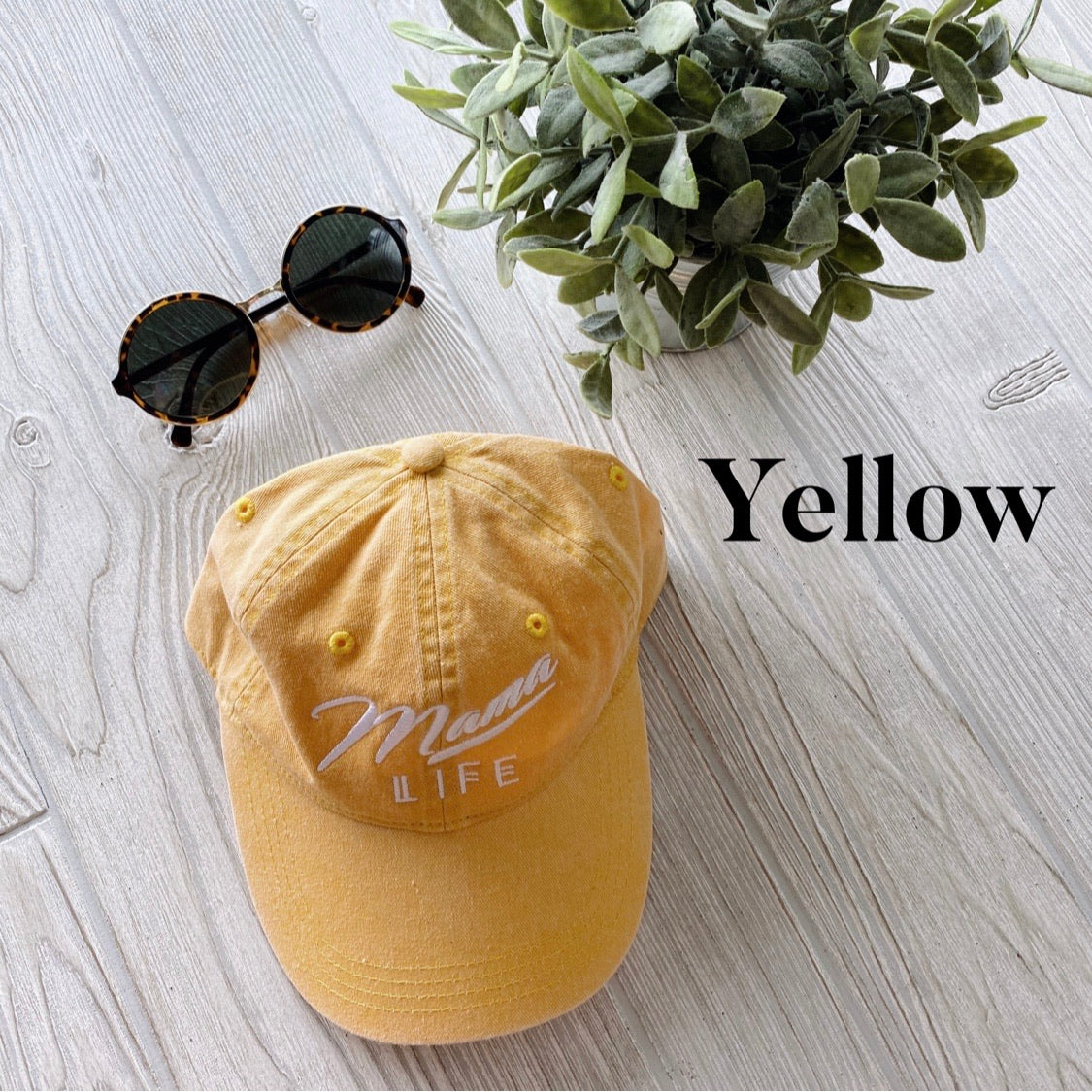 A yellow Mama Life Hat • NEW Colors! embroidered canvas cap sits on a light wood surface next to round sunglasses and a leafy plant. “Yellow” is written in bold black letters on the image’s right side.