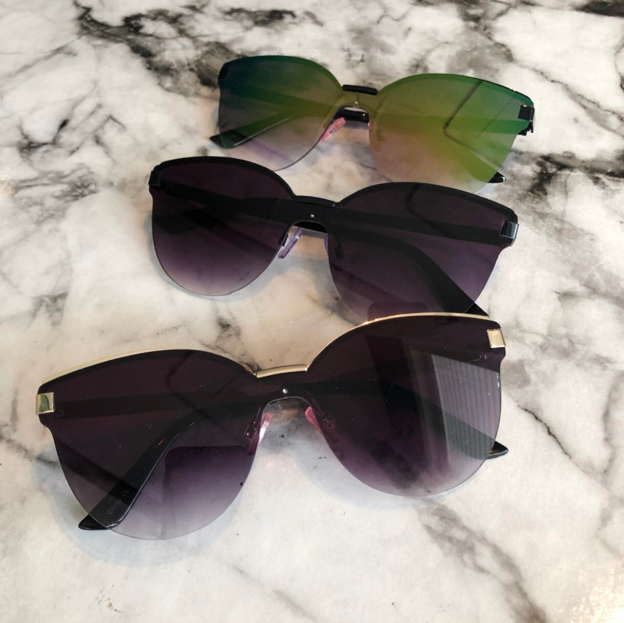 Three pairs of Kim Sunglasses • Adult with UV400 protection and gradient lenses are displayed on white marble. The top pair has green-tinted lenses; the two below feature dark purple lenses. All sunglasses are oversized adult sizes.