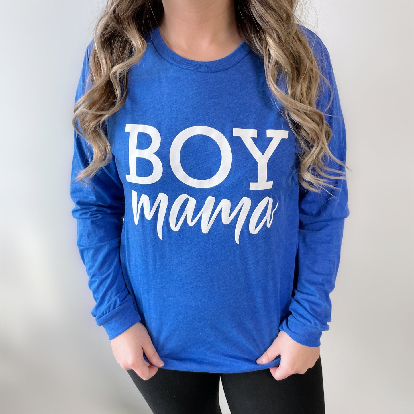 A person with long wavy hair wears the Boy Mama • Royal Long Sleeve shirt, featuring bold white BOY mama lettering, standing against a plain light background.