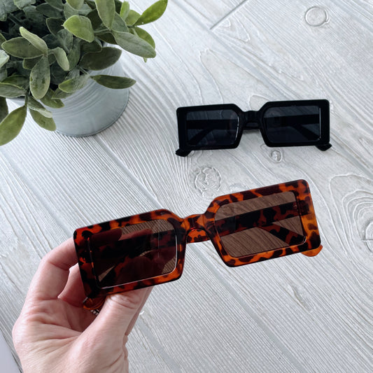 A hand holds Impulse Sunglasses • Adult in square tortoiseshell style with UV400 protection above a light wood surface; a black adult pair and a potted green plant are blurred in the background.