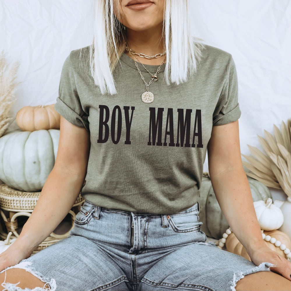 A Proud Boy Mama sits cross-legged in an olive BOY MAMA • Olive Tee with bold black lettering, denim shorts, layered necklaces, and light blonde hair. Pale pumpkins and neutral decor add a cozy touch to the scene.