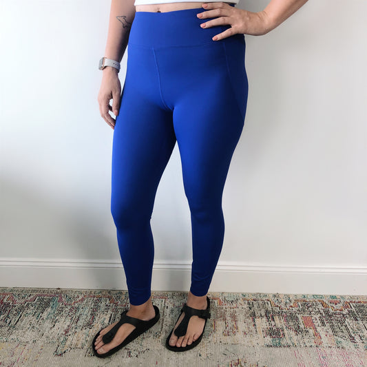 A person wears Billie Legging 6209 in bright blue, paired with black sandals and a wristwatch, standing against a white wall on a multicolored rug with one hand on their hip.