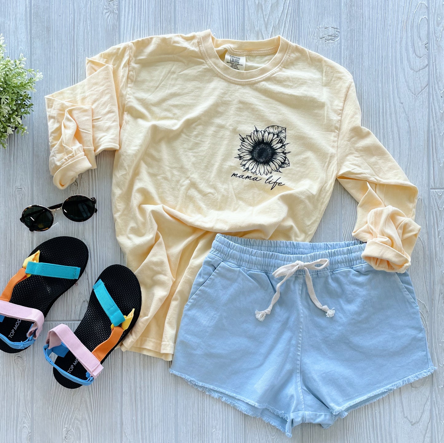 A flat lay of a summer outfit: Mama Life • Sunflower Long Sleeve (light yellow with sunflower print), light blue drawstring shorts, black sandals with colorful straps, and black sunglasses.