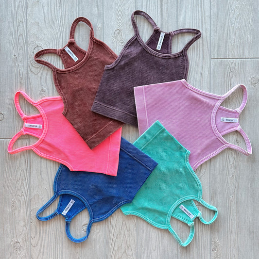 Six Luna Vintage Crop tops from the Tampa Collection in pink, brown, purple, green, blue, and coral are arranged in a circle on a light wood floor. Each features ribbed fabric, a high neck racerback design, and an inner collar tag.