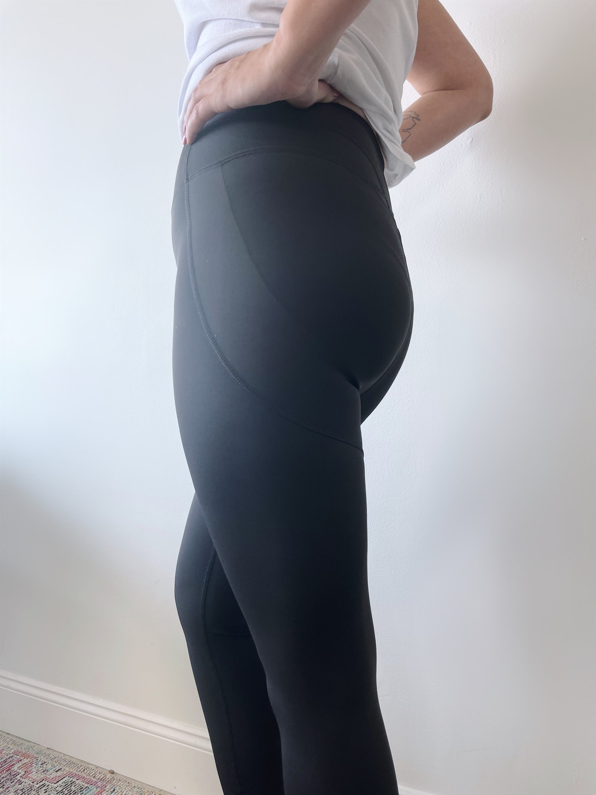A person stands sideways against a white wall, wearing a white top and the Billie Legging 6209 in black, which features tummy control. One hand rests on their hip.