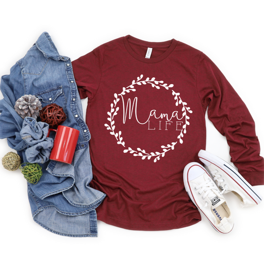 Flat lay of the Mama Wreath • Long Sleeve tee in heathered cardinal with white Mama Life script in a leafy wreath, styled with a denim shirt, white sneakers, decorative balls, scrunchies, and a red cup.