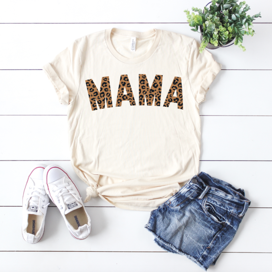 The MAMA Cheetah • Cream Tee is a 100% cotton, cream-colored shirt featuring bold leopard print lettering, styled with denim shorts and white sneakers beside a small green potted plant on a white wooden surface.