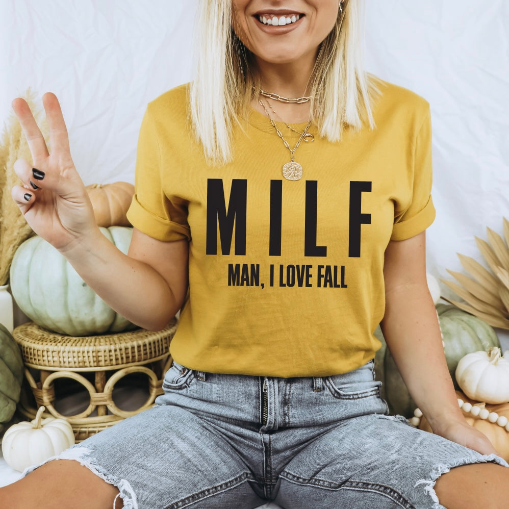 A smiling woman in the MILF • Mustard Tee, layered necklaces, and denim shorts sits among white and green pumpkins, flashing a peace sign.